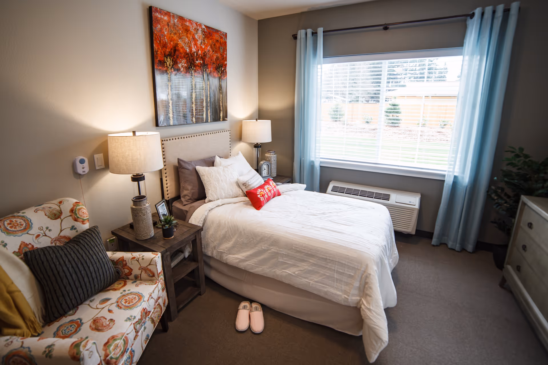 A cozy bedroom with a neatly made bed featuring white bedding and multiple pillows, including a red decorative pillow. There are two bedside tables with lamps, a floral patterned armchair with cushions, a large window with light blue curtains, and a dresser. A pair of pink slippers is placed on the floor next to the bed. A colorful painting of red trees hangs above the bed.