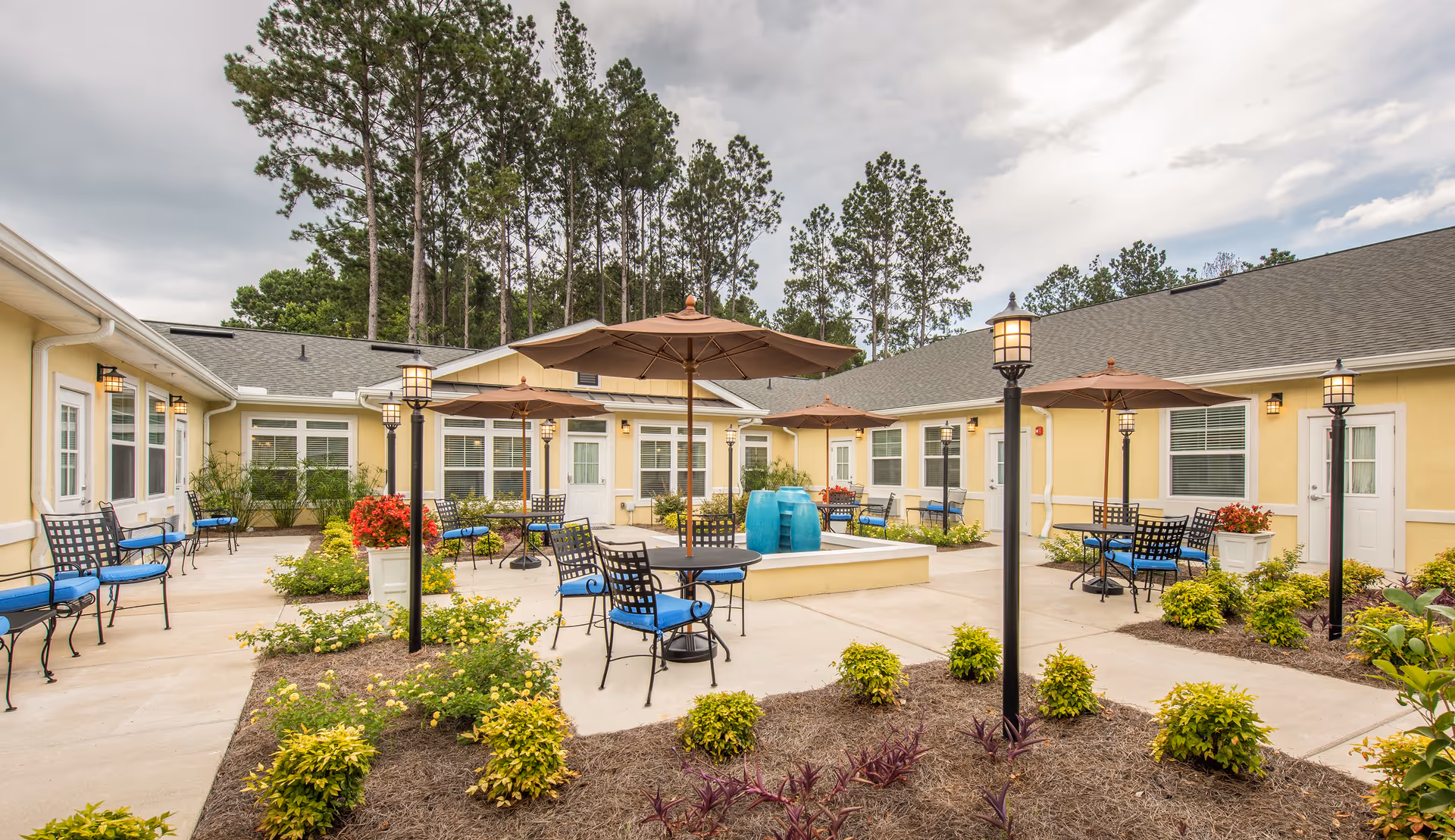 Outdoor courtyard area at The Addison of West Ashley featuring multiple tables with umbrellas and blue cushioned chairs, surrounded by landscaped plants and bushes. The courtyard is enclosed by a single-story yellow building with white trim and multiple windows and doors. Tall pine trees are visible in the background under a partly cloudy sky.