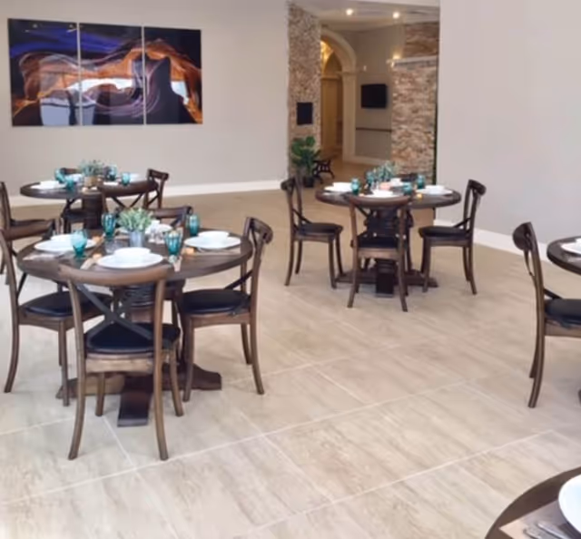 A dining area with round wooden tables set with white plates, teal drinking glasses, and small green plants as centerpieces. The room has light-colored tiled flooring, neutral walls, and a modern abstract painting on one wall. There is a stone accent wall and an arched doorway in the background.