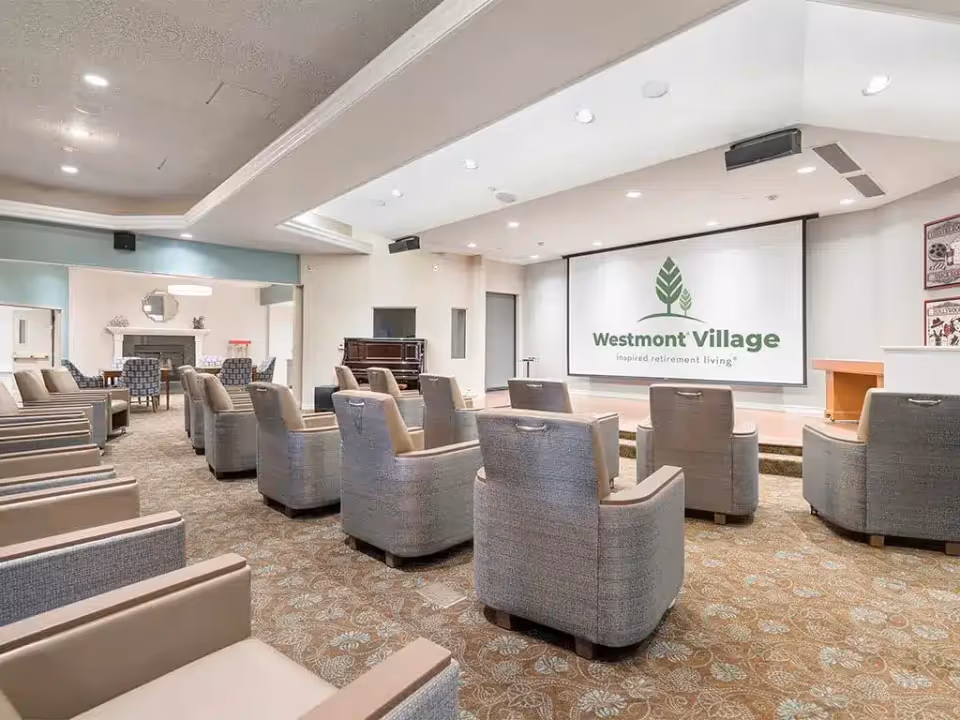 A senior living community auditorium with rows of upholstered chairs facing a large screen displaying the Westmont Village logo.