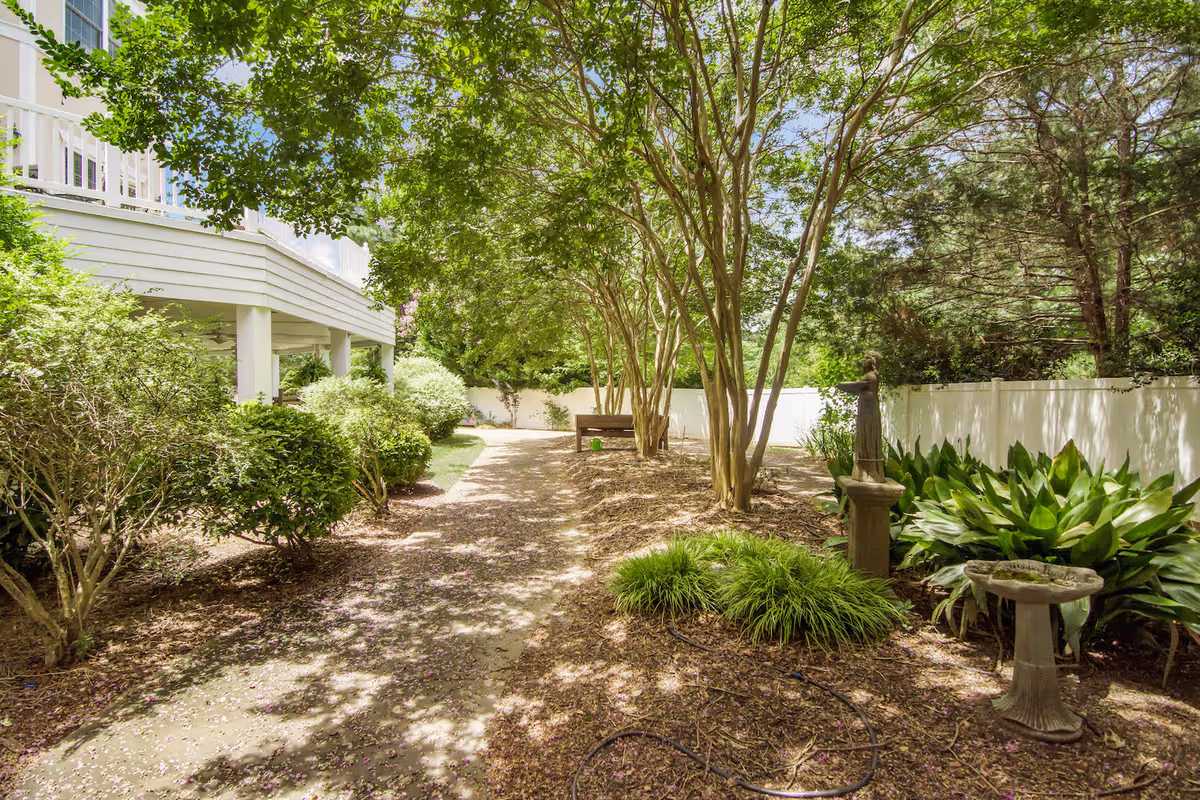 A shaded garden pathway lined with trees, bushes, and plants next to a white building with a porch. There is a wooden bench along the path and a birdbath and small fountain on the right side near a white fence.