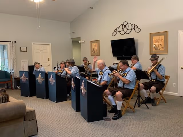 A group of elderly men dressed in traditional Bavarian attire, including hats and lederhosen, are seated in a living room playing musical instruments such as trumpets and trombones. The room has beige carpet, light-colored walls, a wall-mounted TV, framed artwork, and a decorative wall piece above the TV. There is a couch and chairs visible in the foreground and background.