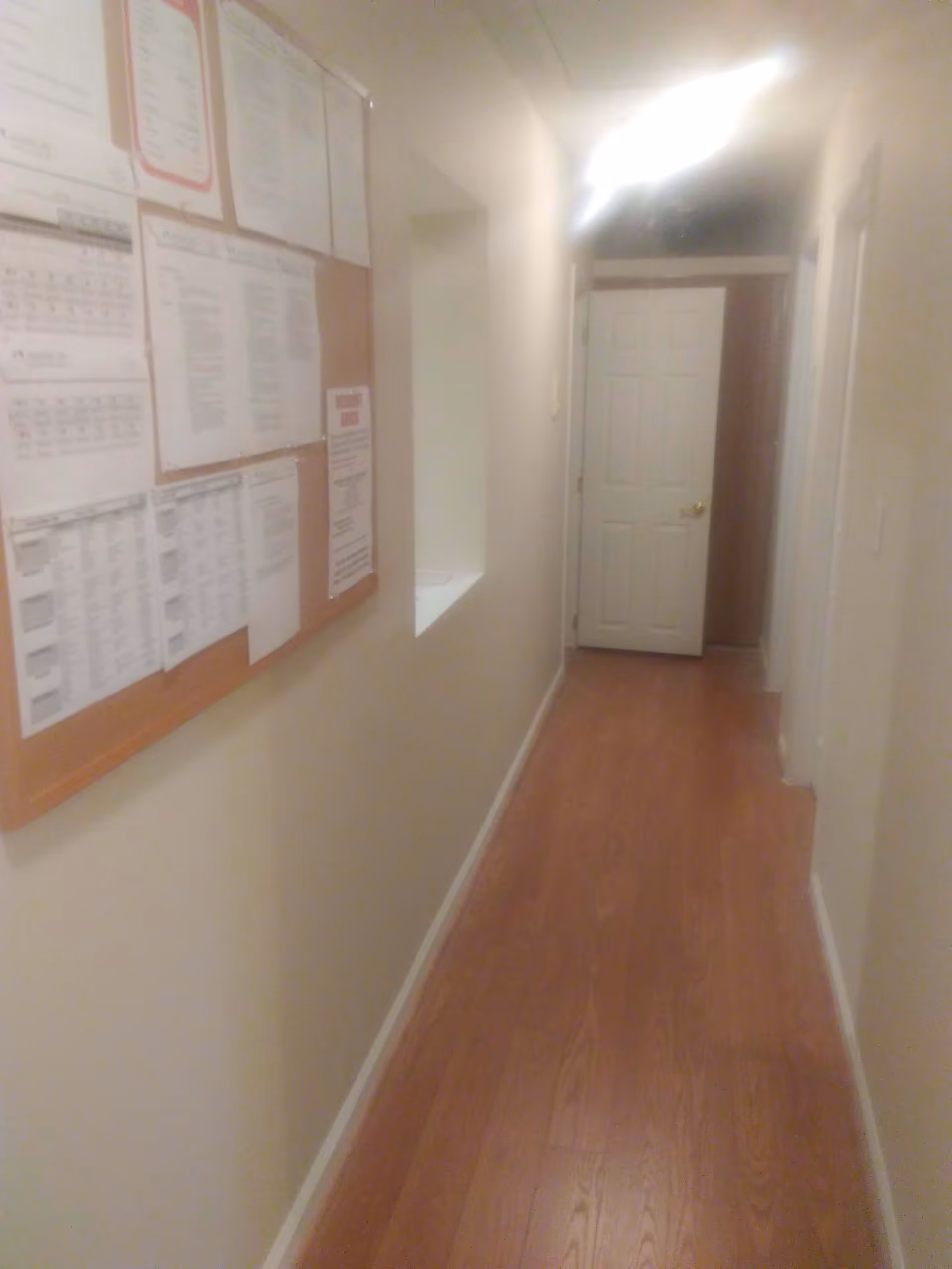 A narrow hallway with wooden flooring and beige walls. On the left wall, there is a bulletin board with various papers and notices pinned to it. At the end of the hallway, there is a white door with a golden handle. The hallway is lit by a ceiling light.