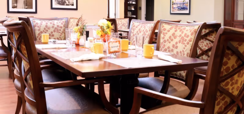 A dining room table set with placemats, yellow mugs, glasses, napkins, and small flower arrangements. The chairs around the table have wooden frames and patterned upholstery. The room has framed pictures on the walls and a wooden floor.