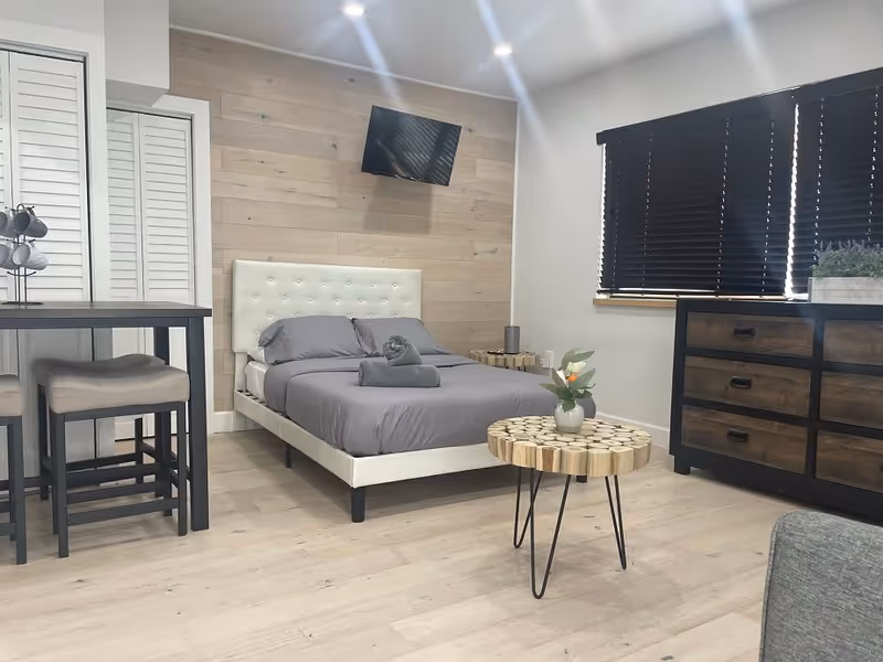 A modern bedroom with a white upholstered bed frame and gray bedding, a small wooden side table with a vase of flowers, a wooden dresser with black handles, a wall-mounted TV, and a small dining table with two cushioned stools. The room has light wood flooring, a wooden accent wall behind the bed, and black window blinds.