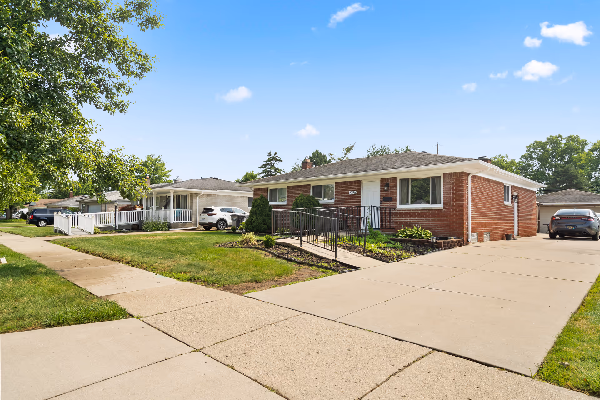 Exterior view of a single-story brick house with a concrete driveway and a wheelchair ramp leading to the front door. The house is surrounded by a well-maintained lawn and neighboring houses are visible along the street under a clear blue sky.