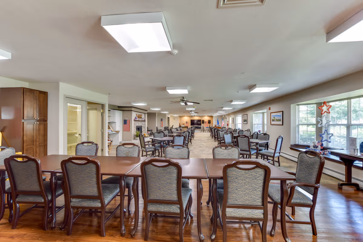 A large, well-lit dining room with multiple tables and chairs arranged in rows. The room has wooden floors, large windows on the right side letting in natural light, and patriotic decorations including American flags and star-shaped ornaments. The far end of the room features a fireplace and a television mounted on the wall.