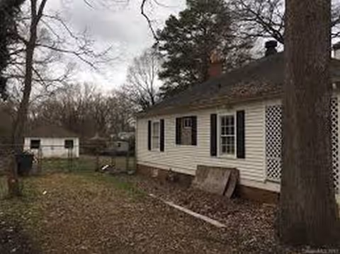 Single-story white building with black shutters and a surrounding yard, trees, and a lattice door.