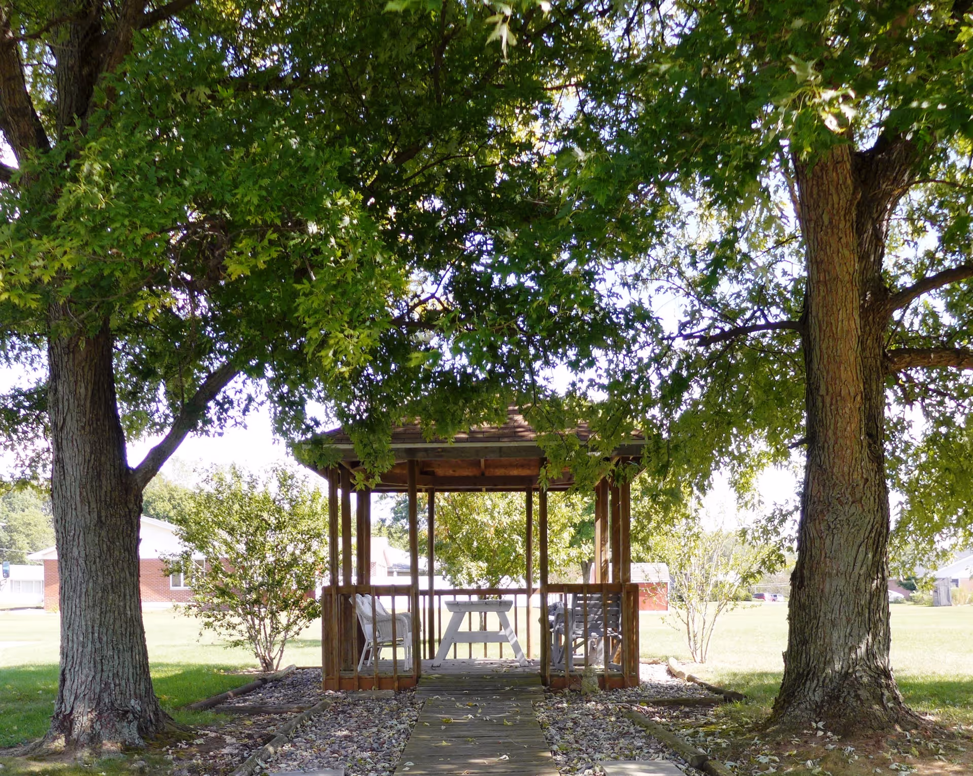 A wooden gazebo with a picnic table sits between two large trees on a grassy lawn.