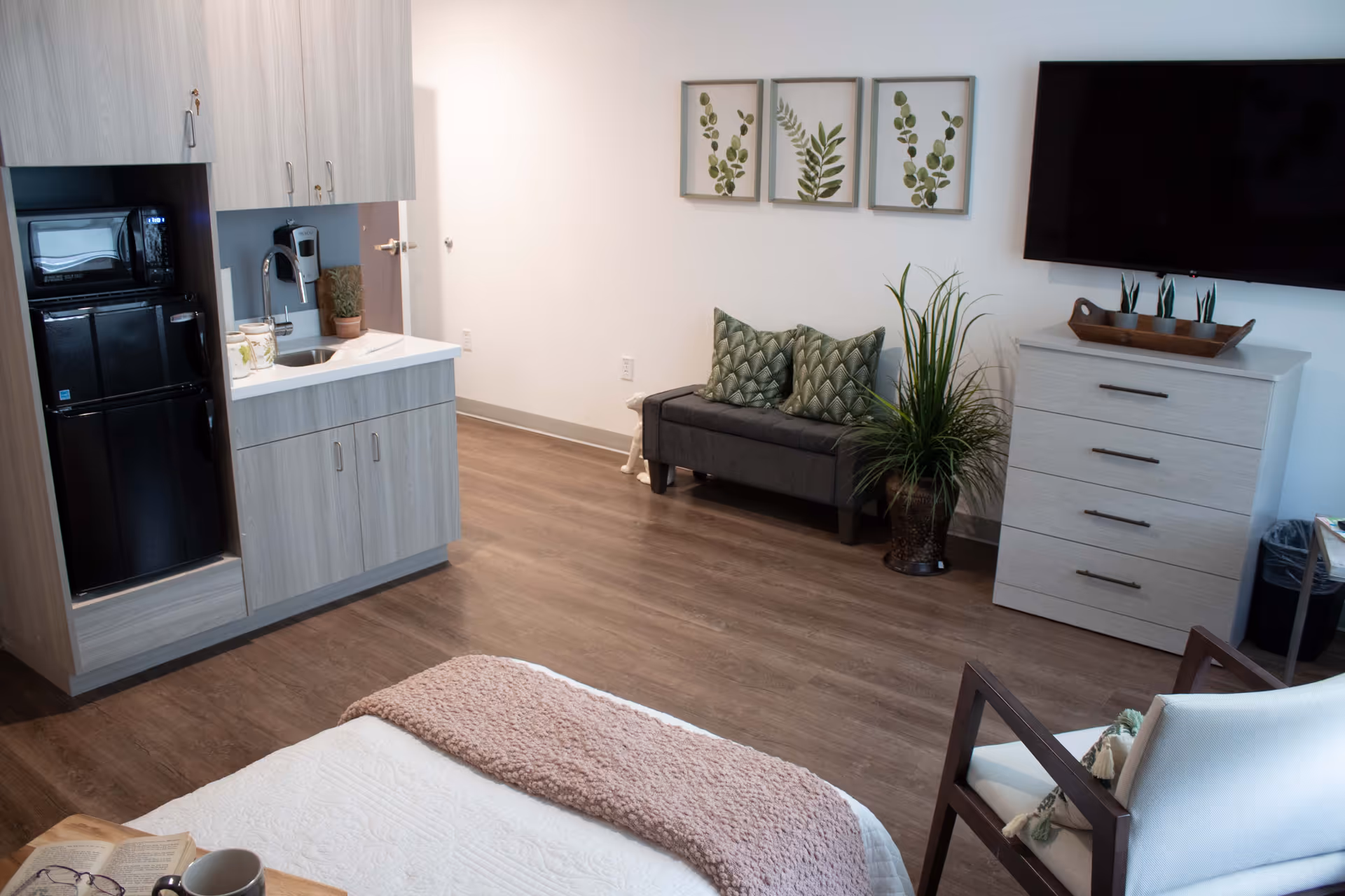 Studio-style senior living apartment with a bed in the foreground, kitchenette on the left, seating area, dresser and wall-mounted TV.