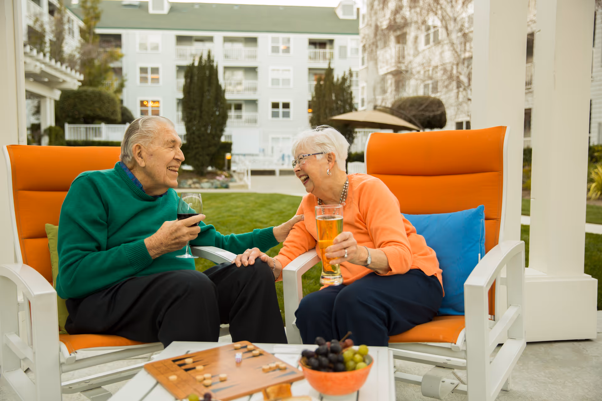 An elderly man and woman sitting outdoors on orange cushioned chairs, smiling and enjoying drinks together. They are seated at a small table with a bowl of grapes and a board game, with a residential building and greenery in the background.