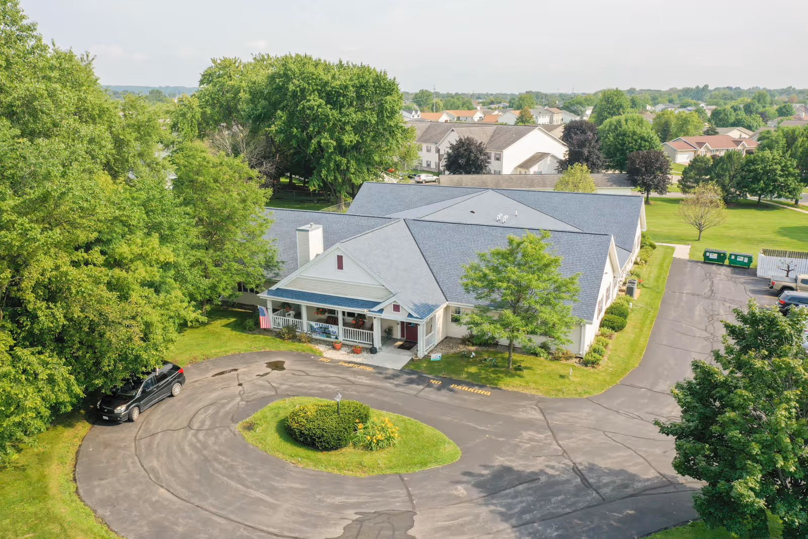 Aerial view of a single-story assisted living building with a circular driveway, front porch, and surrounding trees and lawn.