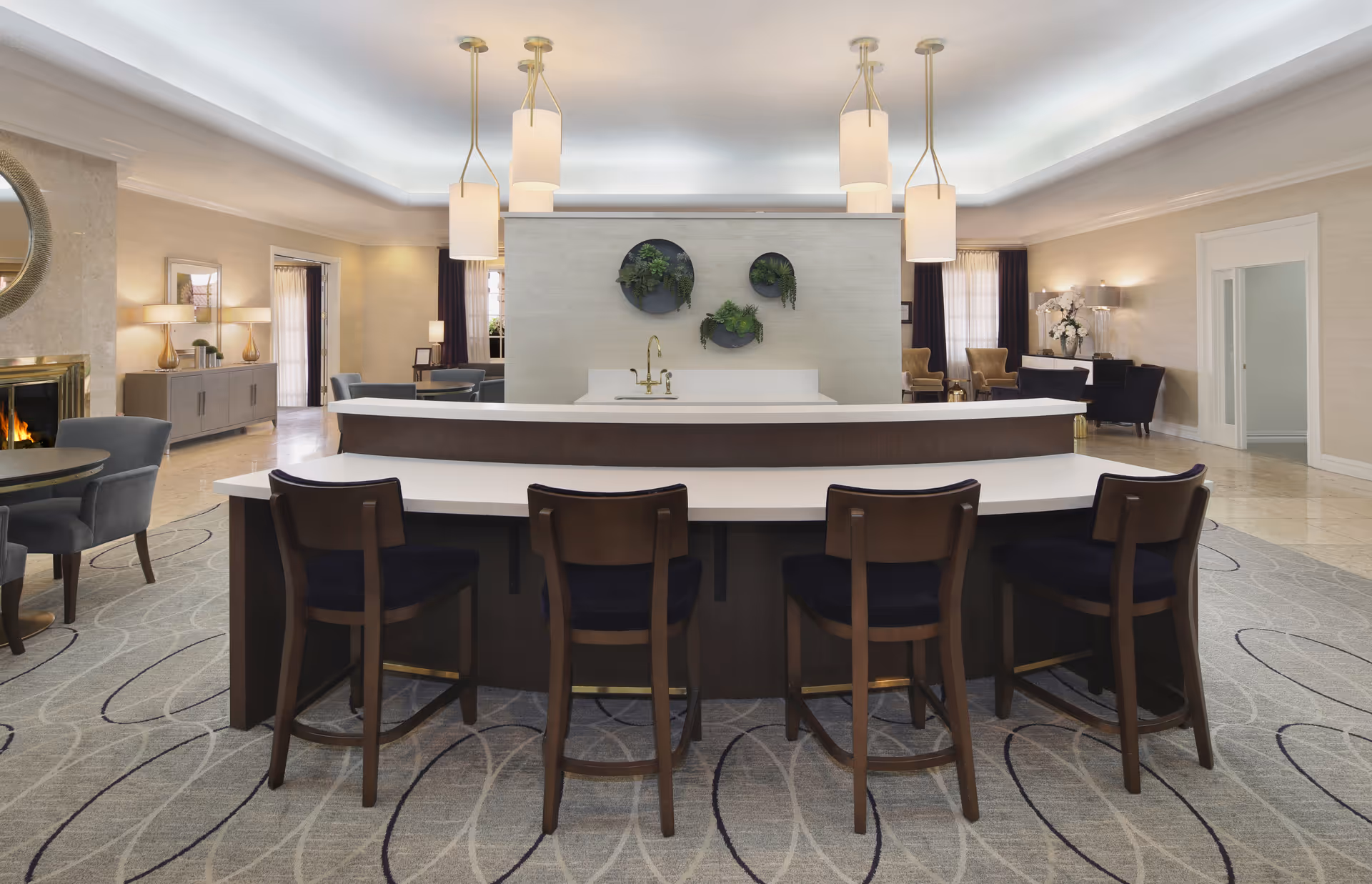 A spacious and elegant common area with a curved white countertop bar and four wooden chairs with dark cushions. Behind the bar is a wall with three circular planters containing green plants. The room features soft lighting with pendant lamps hanging from the ceiling, a fireplace on the left, and several seating areas with armchairs and tables. The floor is covered with a patterned carpet and the walls are light-colored with decorative elements.