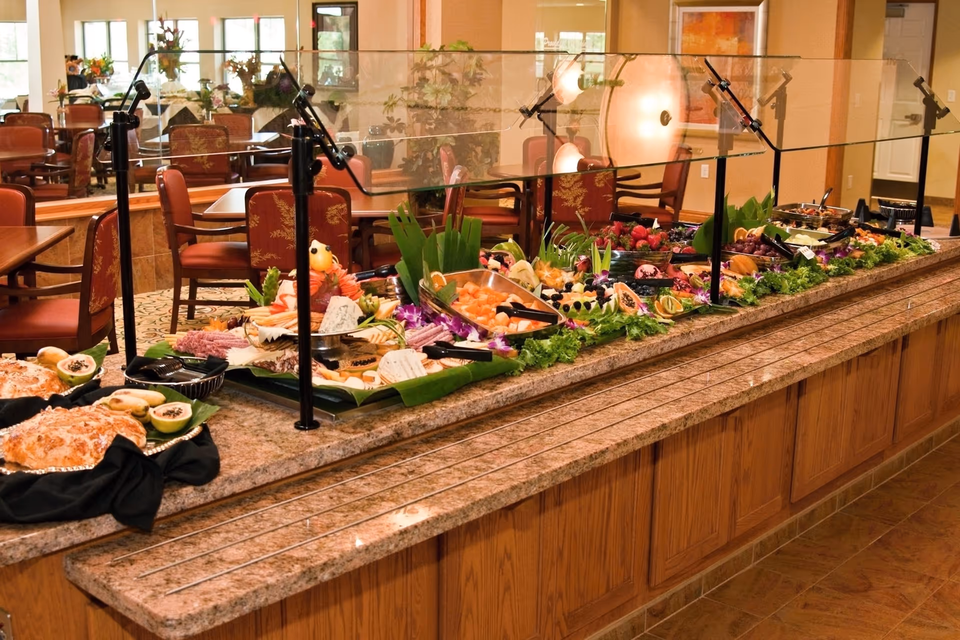 Buffet-style food serving area with a variety of fresh fruits, cheeses, and other dishes displayed on a granite countertop with a glass sneeze guard. In the background, there are dining tables and chairs with red upholstery and floral patterns in a well-lit dining room.