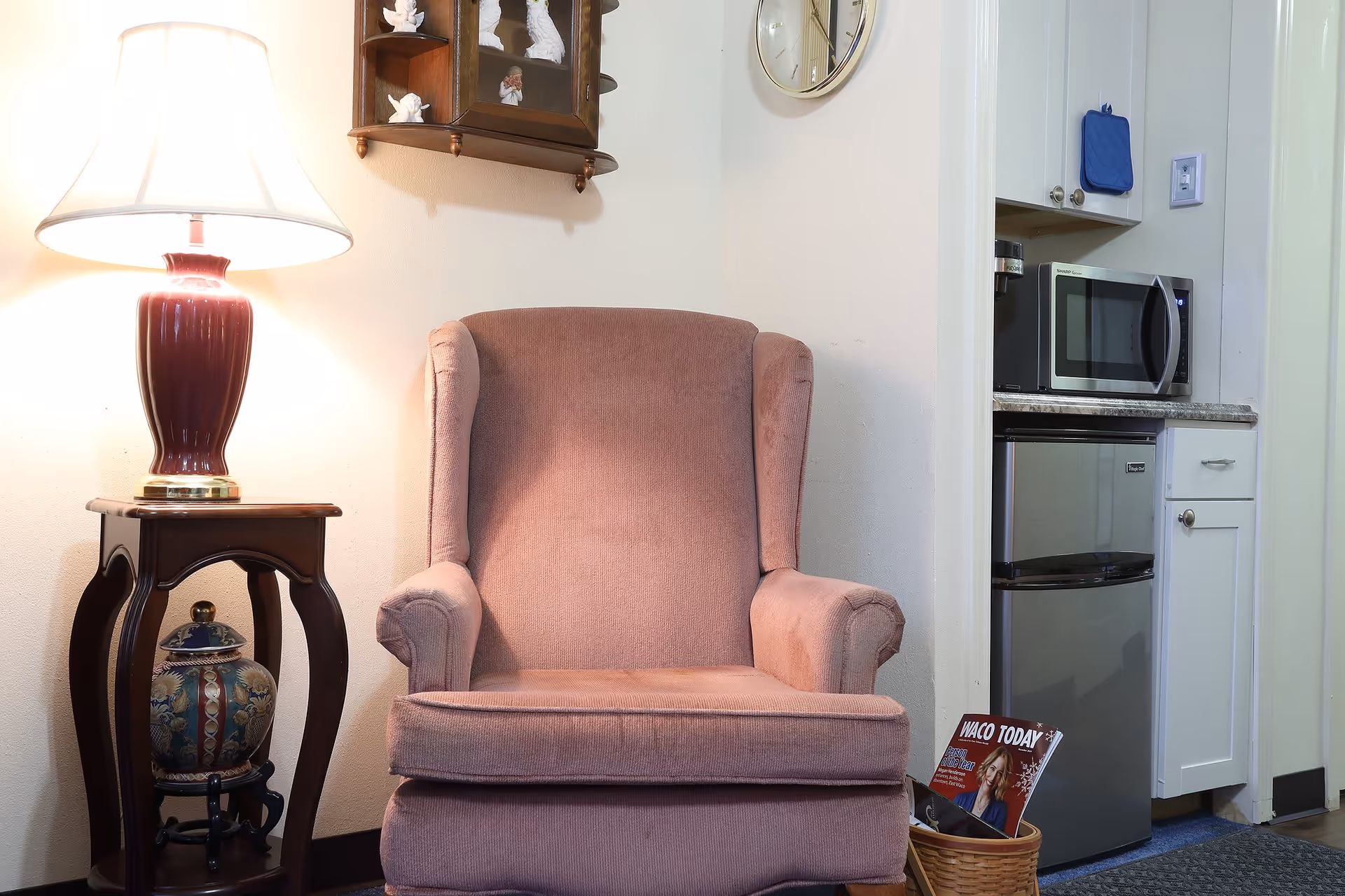 Upholstered armchair next to a side table with a lamp, with a small kitchenette area including a microwave and mini-fridge visible to the right.