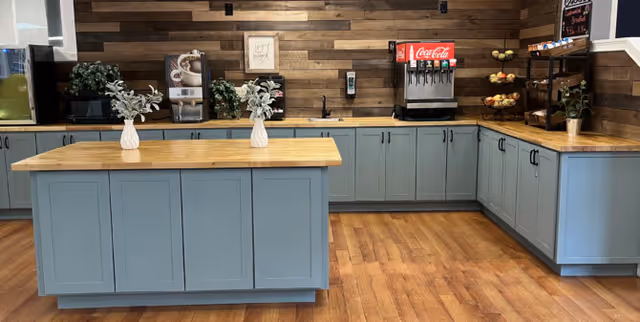 A beverage and snack station area with light blue cabinets and wooden countertops. There is a central island with two small white vases holding green plants. The back wall has a Coca-Cola soda fountain machine, a microwave, a coffee machine, and a tiered fruit basket with apples and bananas. The floor is wooden, and the wall behind the counter is decorated with horizontal wooden panels.