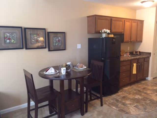 Small kitchenette and dining area with a round table set for two, wooden chairs, a black refrigerator, and wooden cabinets.
