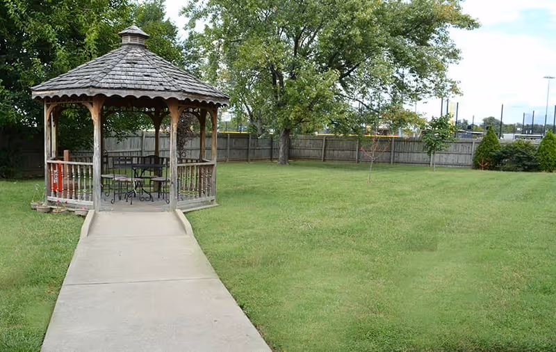 A wooden gazebo with a shingled roof situated at the end of a concrete pathway in a grassy fenced yard with trees and shrubs.
