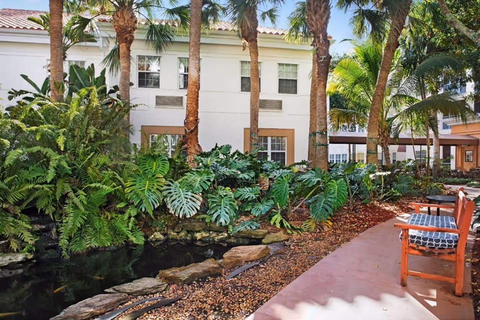 Outdoor garden area at Stratford Court of Boca Pointe featuring lush green plants, palm trees, a small pond with rocks, and a paved walkway with wooden benches and a small table.