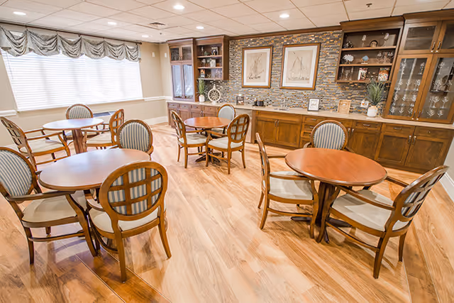 Bright communal dining room with round wooden tables and chairs and built-in wood cabinets with framed artwork on the back wall.