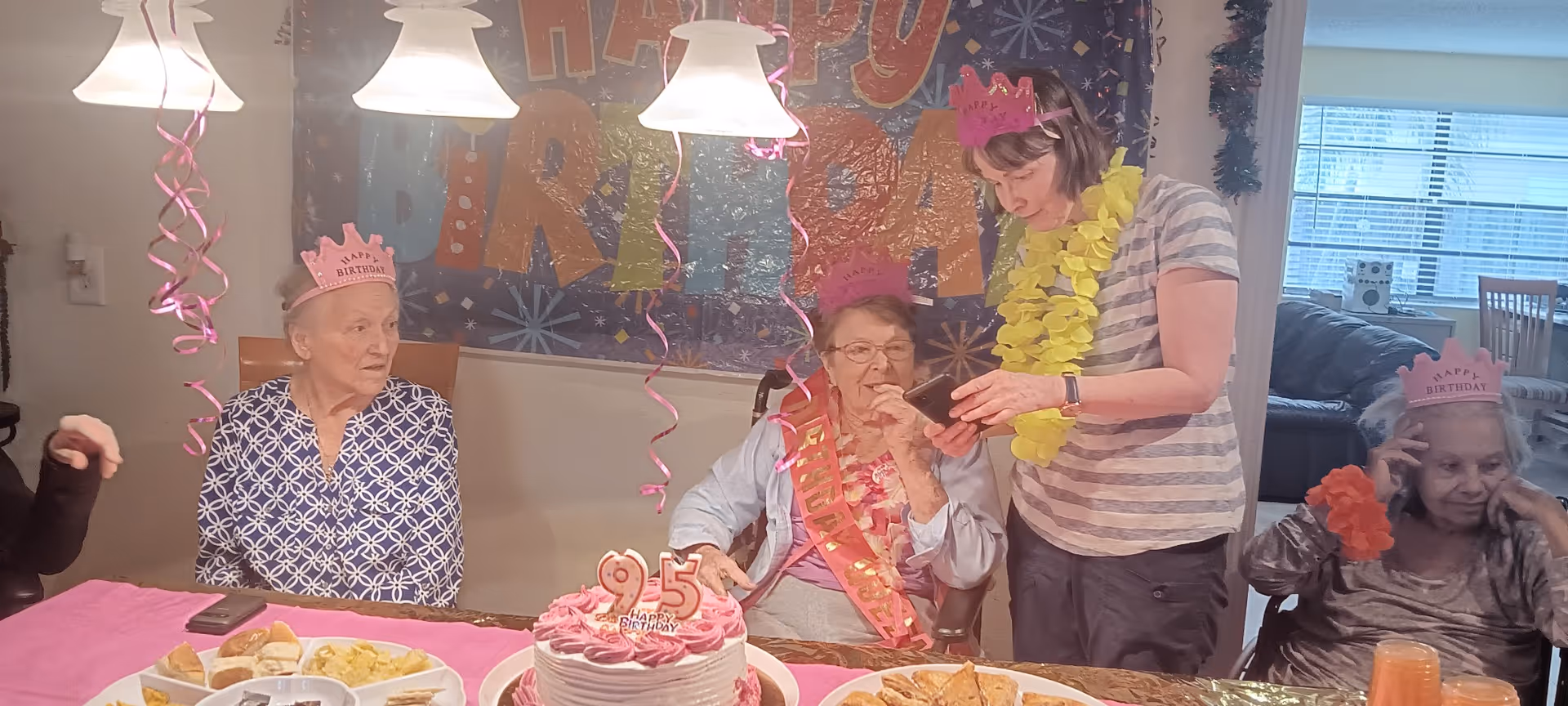 A birthday celebration with elderly women wearing pink 'Happy Birthday' crowns sitting around a table with a pink birthday cake that has a '95' candle on it. One woman is wearing a sash and another woman is standing, showing something on a phone to the woman with the sash. The room is decorated with a 'Happy Birthday' banner and pink hanging streamers.