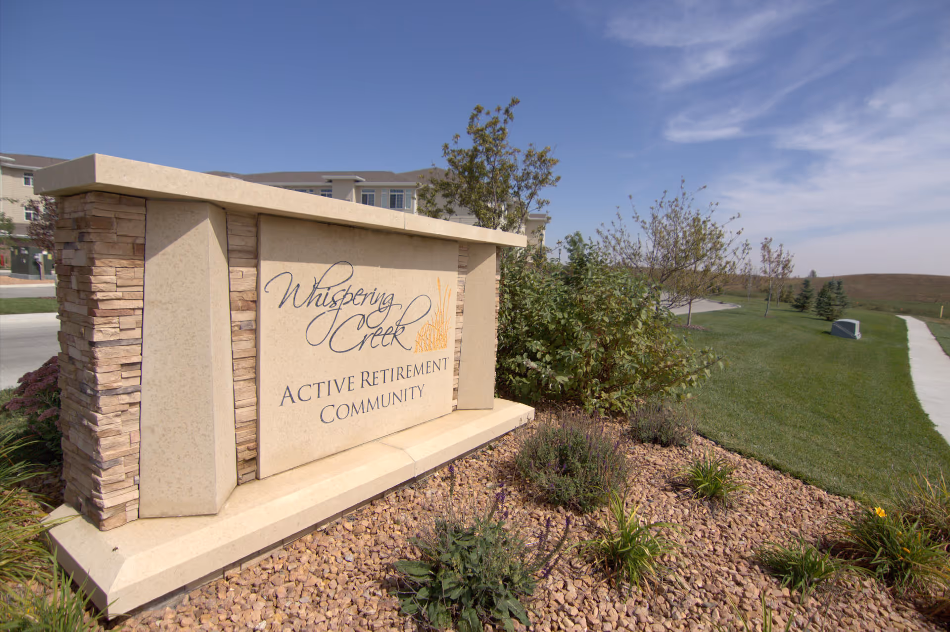 Stone sign at the entrance of Whispering Creek Active Retirement Community with landscaping of rocks, plants, and bushes, and a sidewalk and grassy area extending into the distance under a partly cloudy sky.