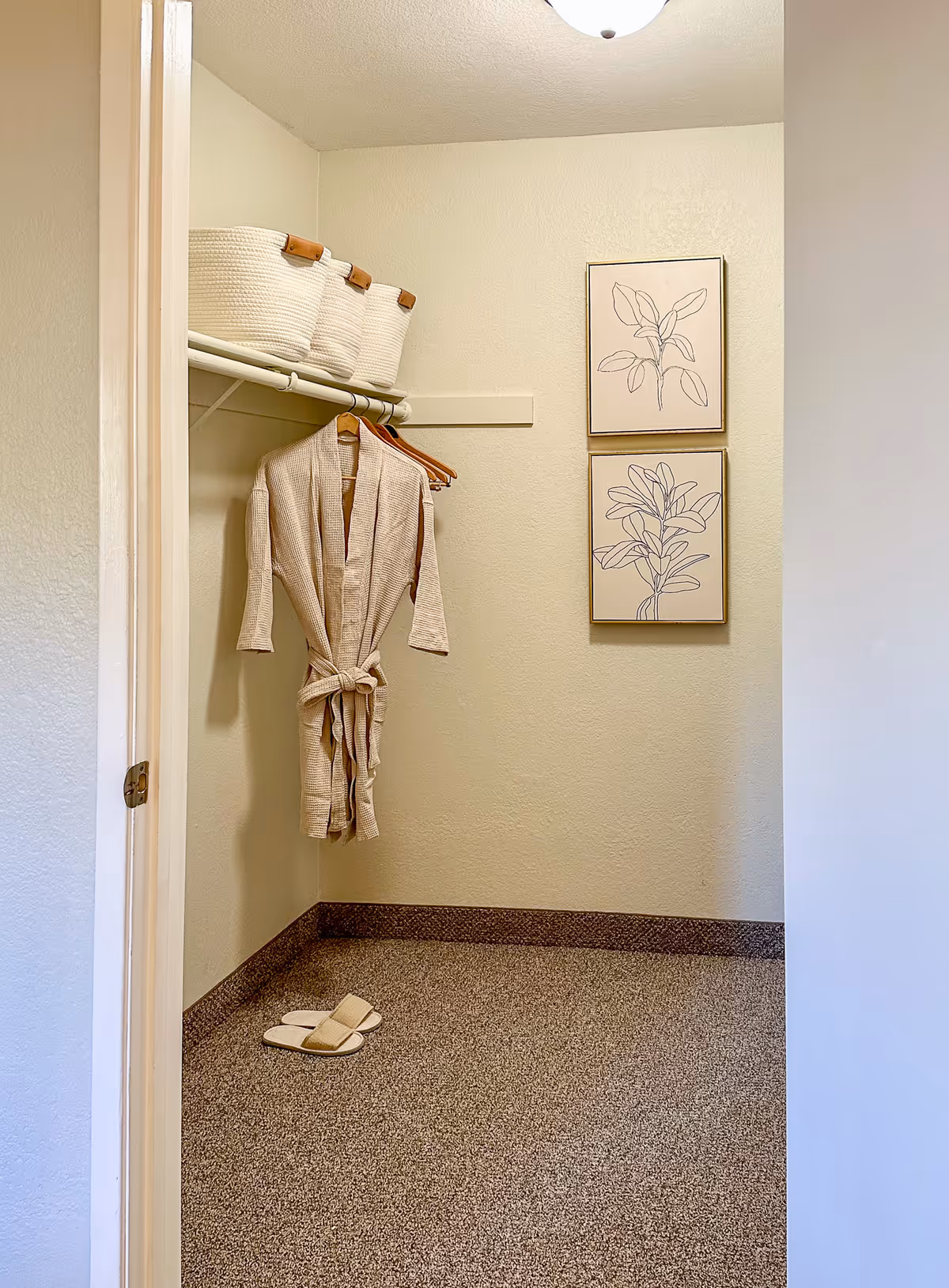 A small closet with a beige robe hanging on a wooden hanger, three white woven baskets on a shelf above, a pair of beige slippers on the floor, and two framed botanical line drawings on the wall.