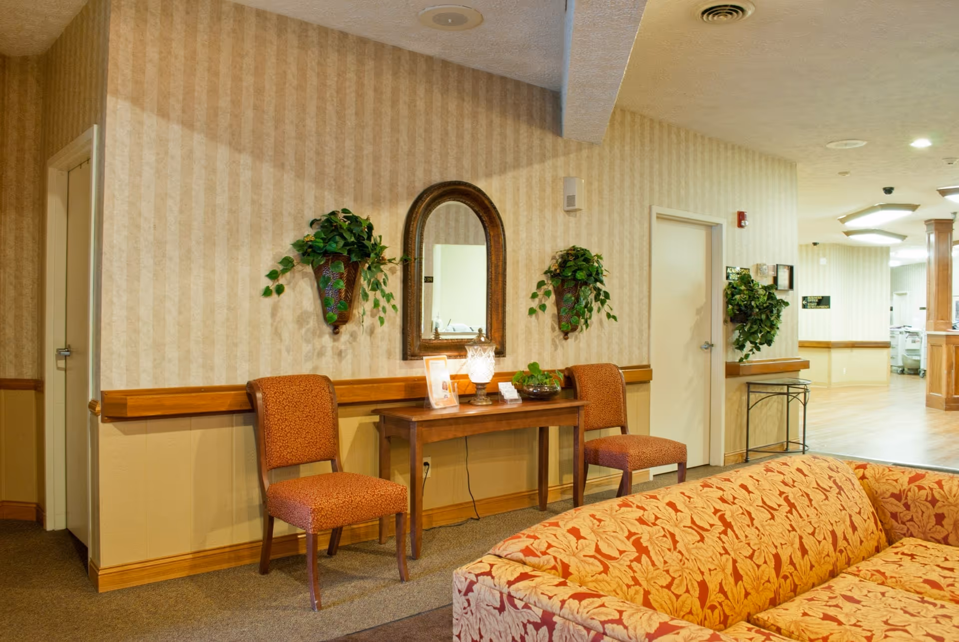 Interior view of a senior living facility lounge area with two orange patterned chairs flanking a wooden table with a lamp, a mirror, and plants on the wall. A patterned sofa is partially visible in the foreground, and a hallway with wood flooring and additional plants is seen in the background.