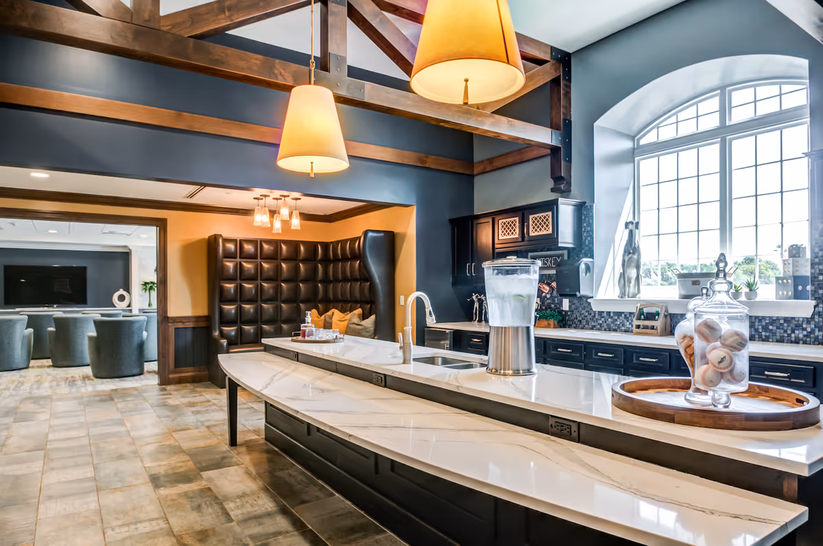 A modern kitchen area with a long marble countertop and a built-in sink. There are two large pendant lights hanging from a wooden beam ceiling. On the counter, there is a glass container with water and a tray holding a glass jar filled with decorative balls. In the background, there is a cozy seating nook with tufted leather upholstery and pillows, and an adjacent room with multiple gray armchairs facing a large flat-screen TV. A large window lets in natural light.