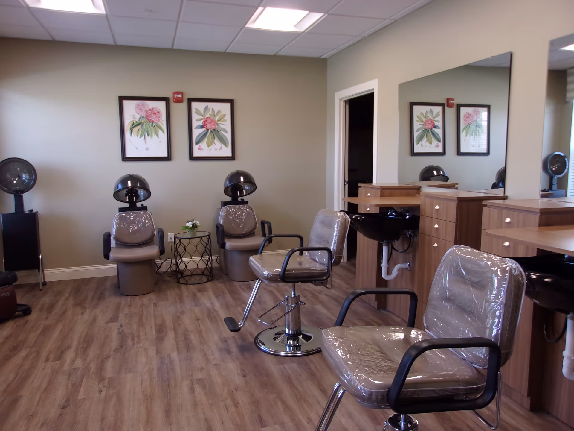 Interior of a salon area in a senior living facility with salon chairs covered in plastic, hair drying stations, wooden cabinetry with sinks, and two framed botanical prints on a beige wall.