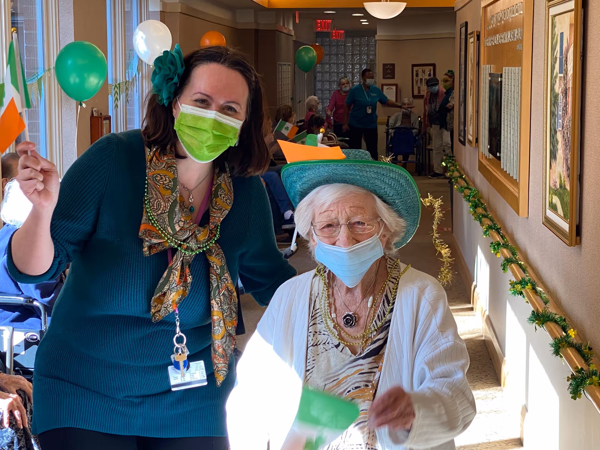 An elderly woman wearing a green hat and a face mask is standing next to a younger woman also wearing a face mask and a green sweater. They are in a decorated hallway with green and orange balloons and garlands, celebrating an event. Other people are visible in the background, some seated and some standing.