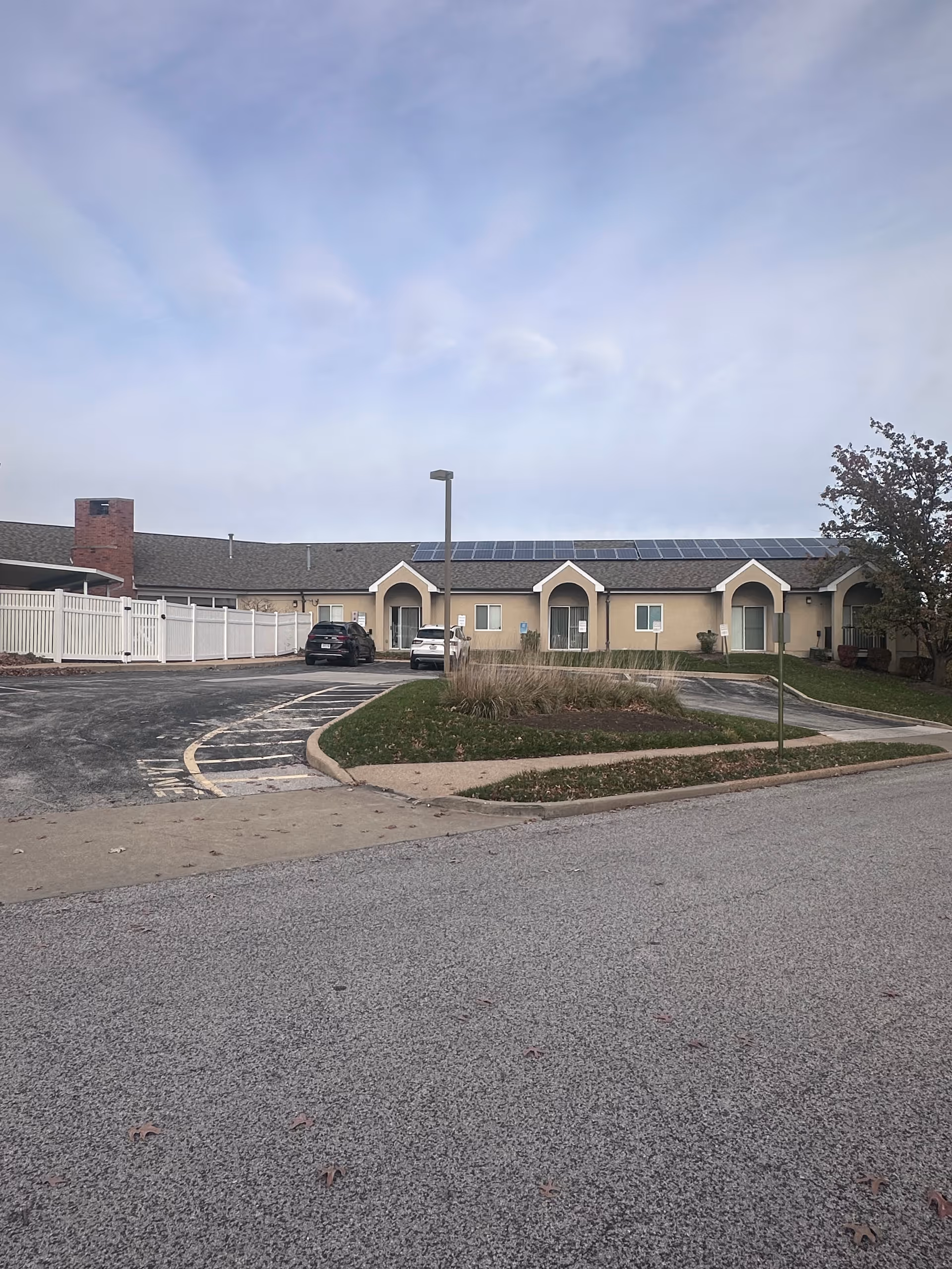 Exterior view of a single-story building with beige walls and a gray shingled roof featuring solar panels. There are three arched entryways, a white fence on the left side, a parking lot with two cars, a street lamp, and some landscaping with grass and bushes. The sky is partly cloudy.