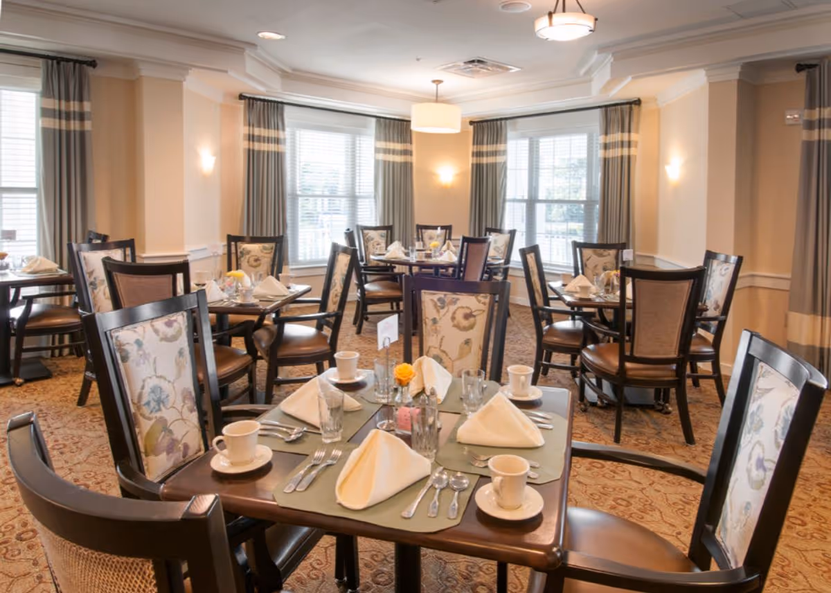 Bright dining room with multiple tables set for a meal and upholstered chairs near large windows.