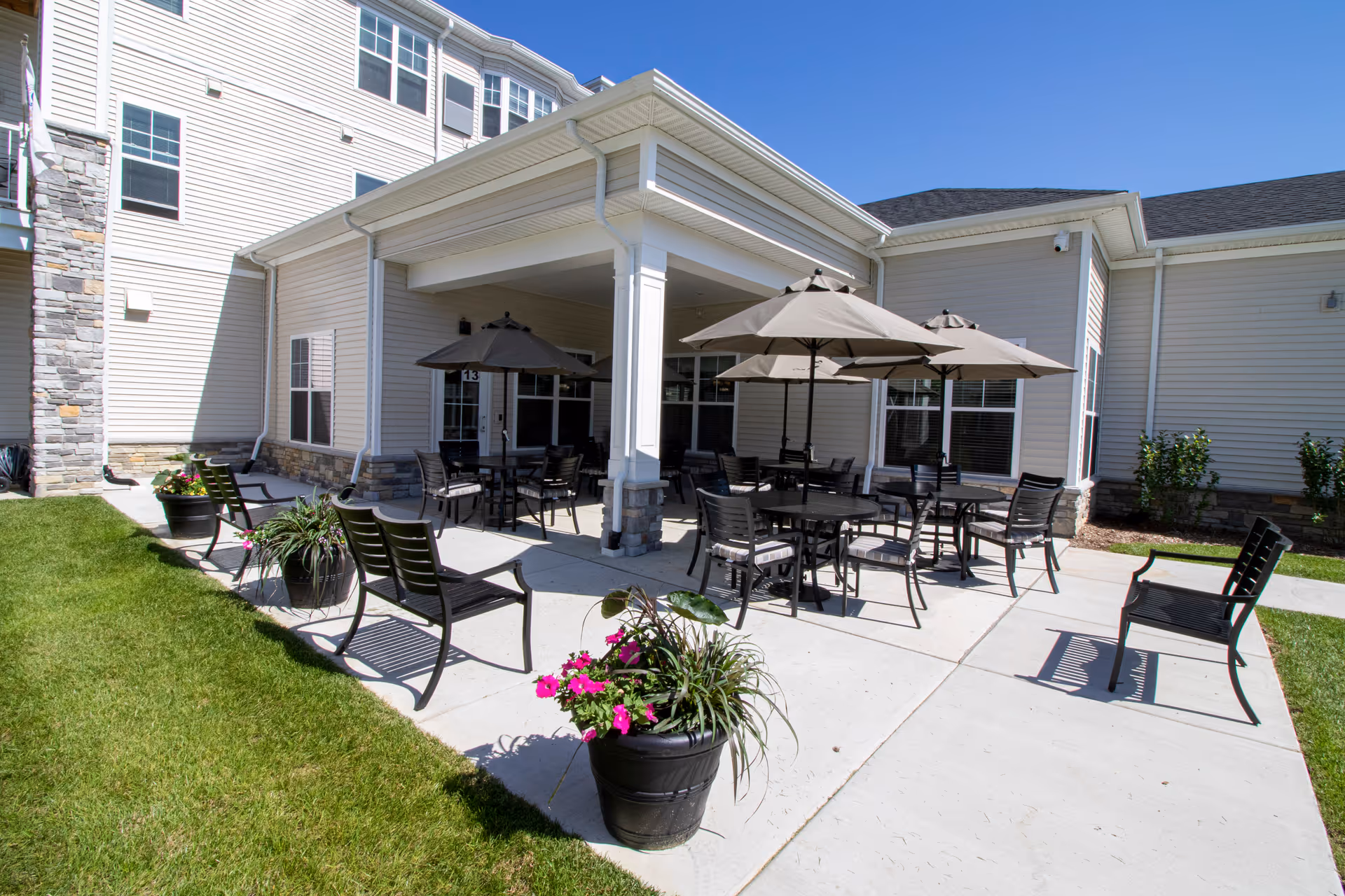 Outdoor patio area at StoryPoint Fairfield with several black metal tables and chairs, each table shaded by large beige umbrellas. The patio is adjacent to a light-colored building with multiple windows. There are also black benches and large flower pots with green plants and purple flowers along the concrete walkway next to a grassy lawn under a clear blue sky.