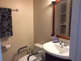 Small bathroom with a toilet equipped with safety rails, a white sink with a wooden framed mirror above it, a towel rack with a dark floral patterned towel, and beige walls.