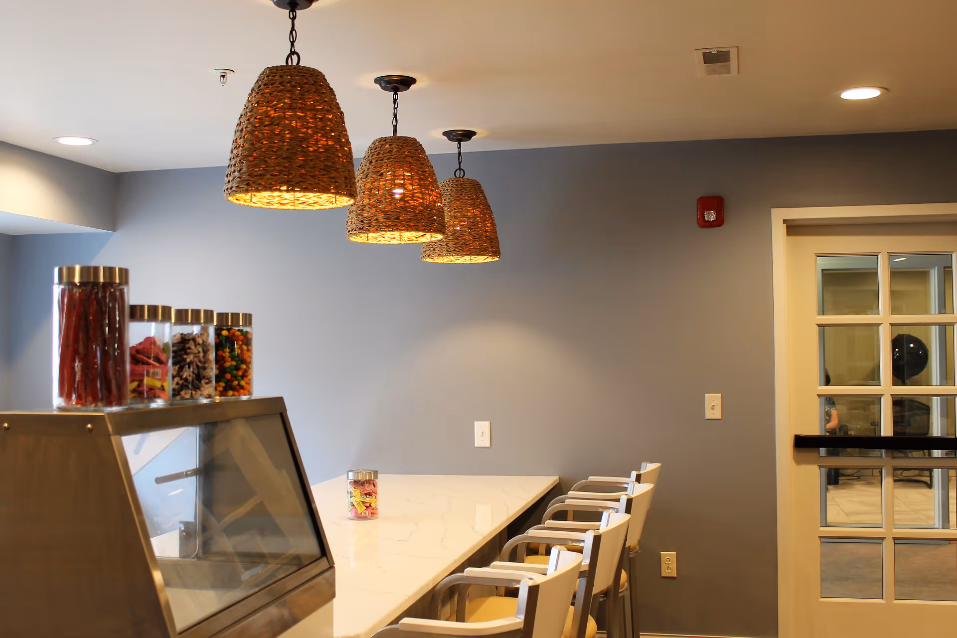 Interior view of a dining area with a long white marble table surrounded by white chairs. Above the table, there are three woven pendant lights hanging from the ceiling. On the left side, there is a display case with jars of colorful candies on top. The walls are painted gray, and there is a glass-paneled door on the right side.