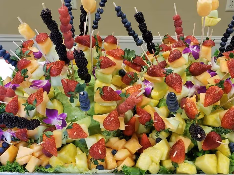 A colorful arrangement of fresh fruit skewers featuring strawberries, blueberries, blackberries, cantaloupe, honeydew melon, and pineapple, garnished with purple edible flowers and green leafy lettuce.