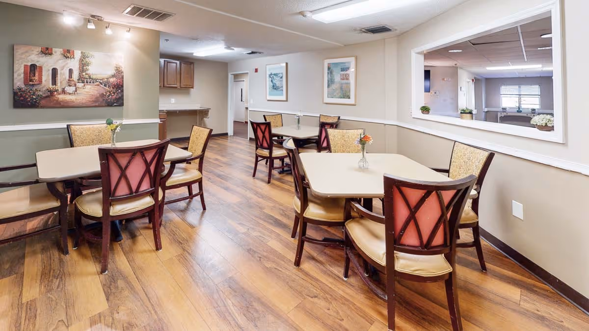 Communal dining area with multiple tables and chairs on hardwood floors, wall artwork, and a pass-through window.