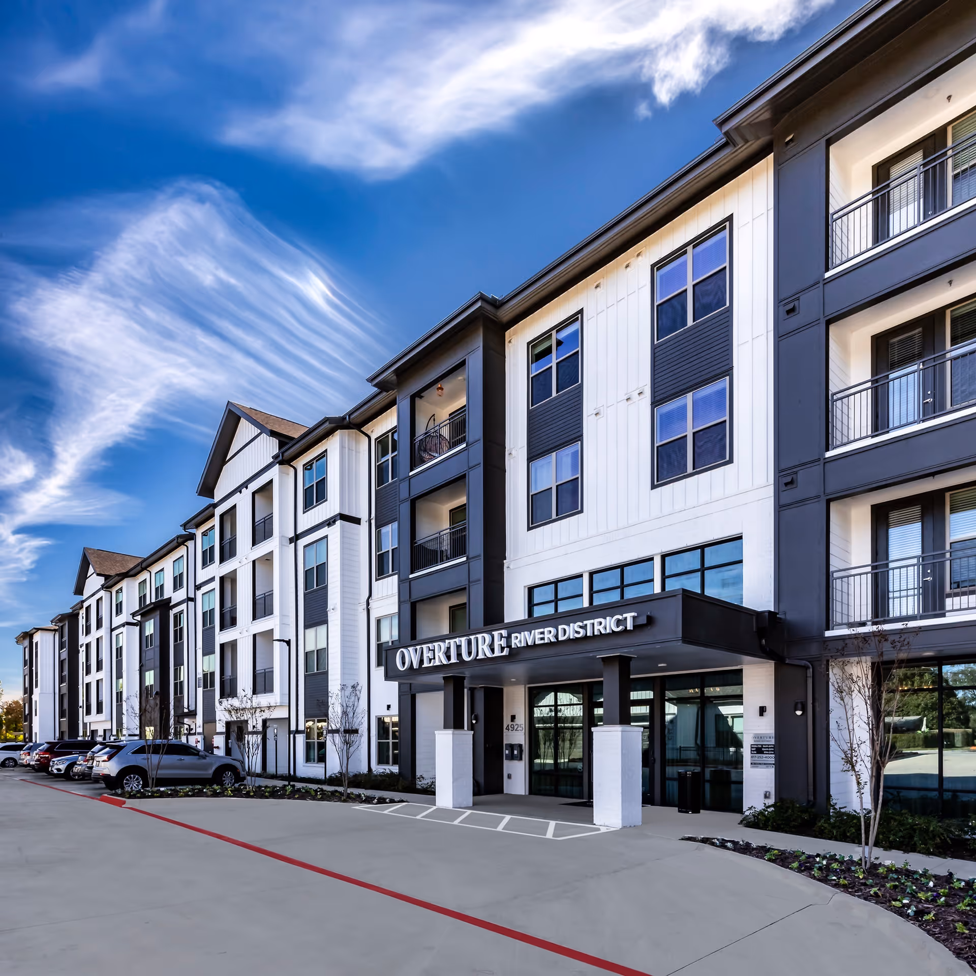 Exterior view of a modern multi-story building with a sign reading 'Overture River District' above the entrance. Several cars are parked along the driveway, and the sky is clear with some wispy clouds.