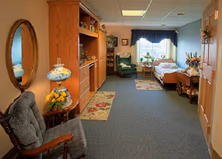 Furnished senior resident room with a bed, seating area, wooden cabinets, and decorative accents.