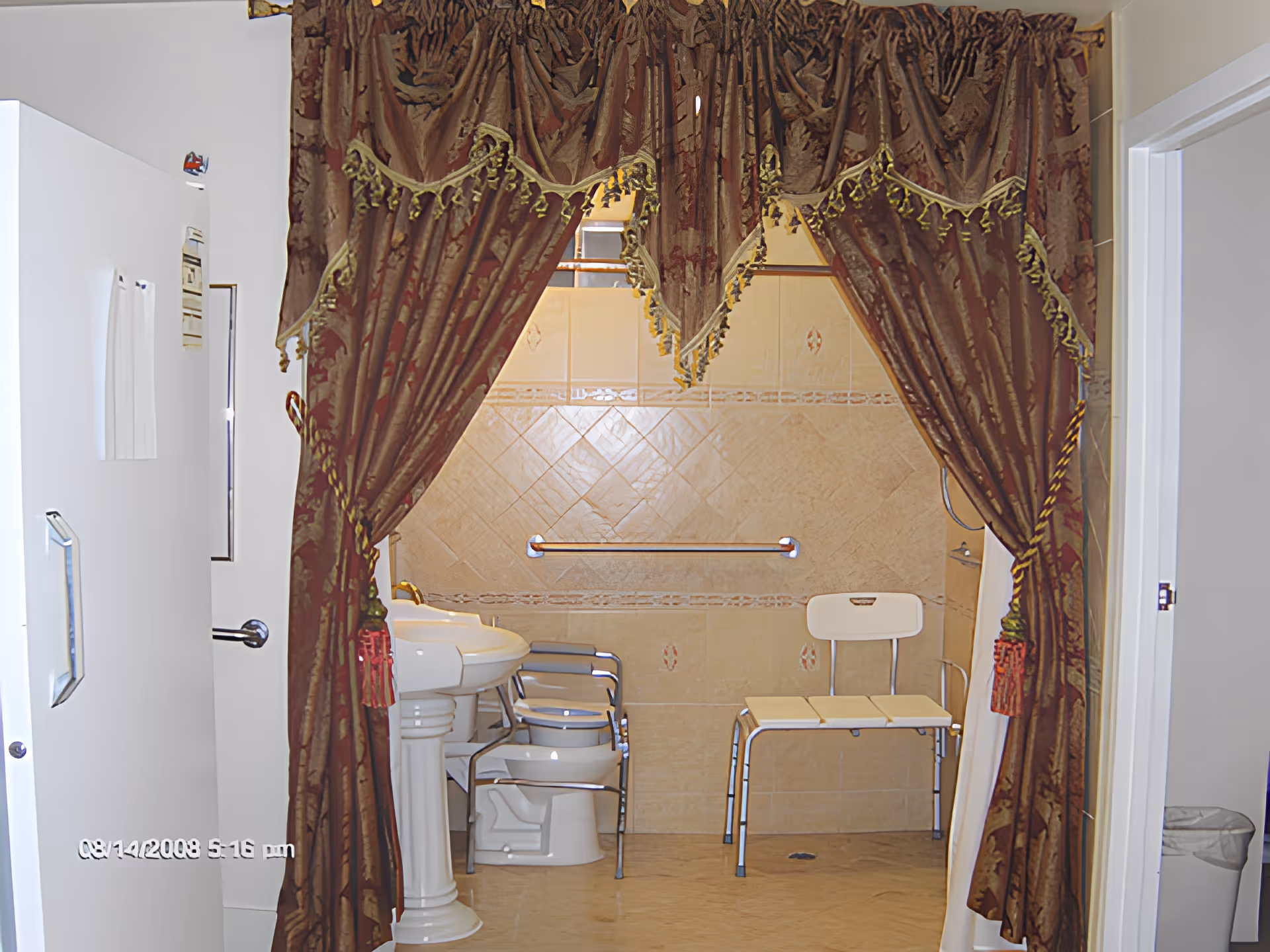 Accessible bathroom with heavy decorative curtains framing a walk-in shower area containing a shower chair, grab bar, pedestal sink and commode.