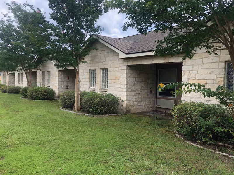 Single-story stone nursing facility exterior showing windows, shrubs, trees, and a grassy lawn.