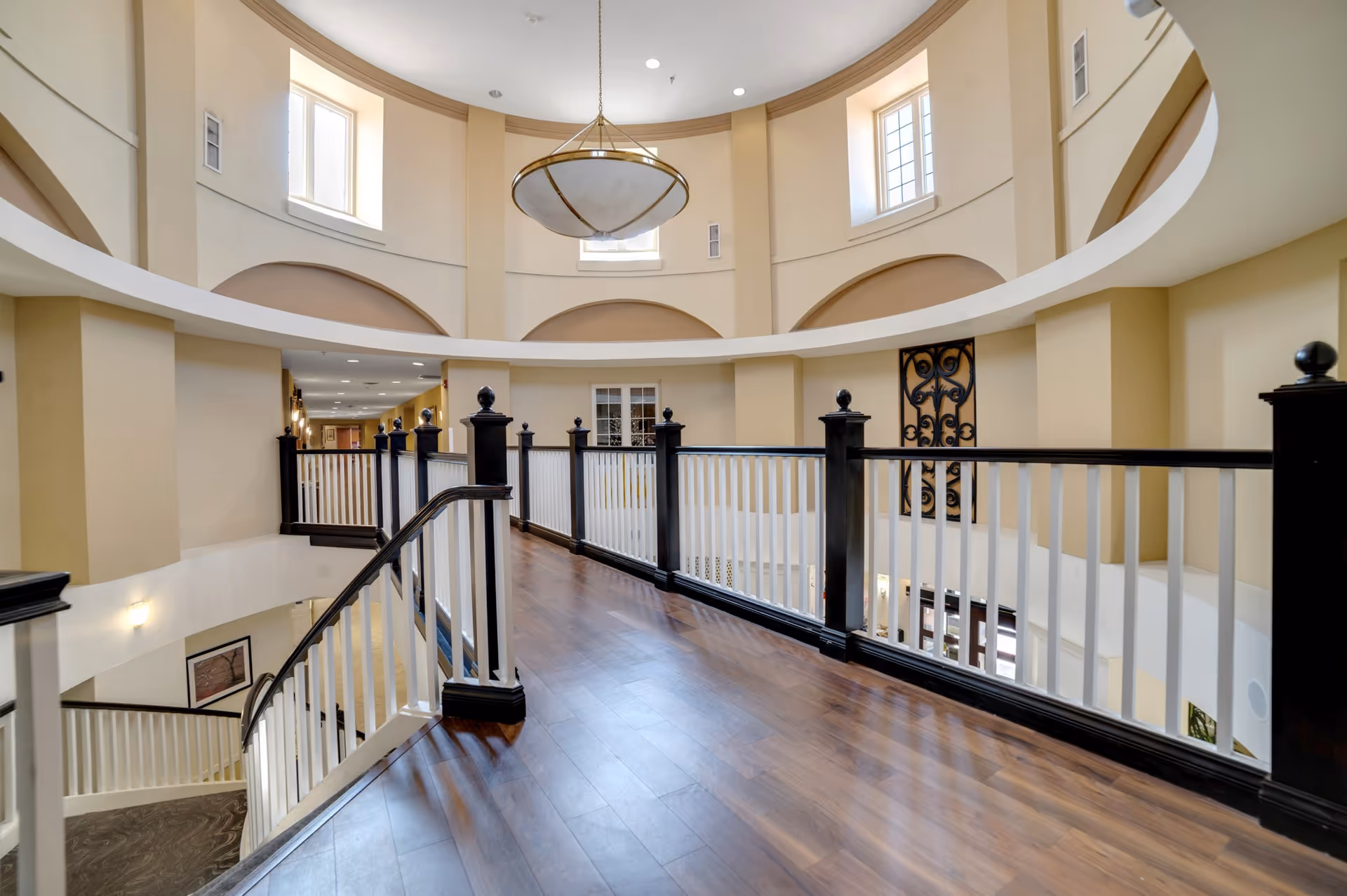 Interior view of a senior living facility hallway with wooden flooring, white and black railing, beige walls, and a large circular chandelier hanging from the ceiling. The hallway overlooks a lower level with a curved staircase and multiple windows allowing natural light.