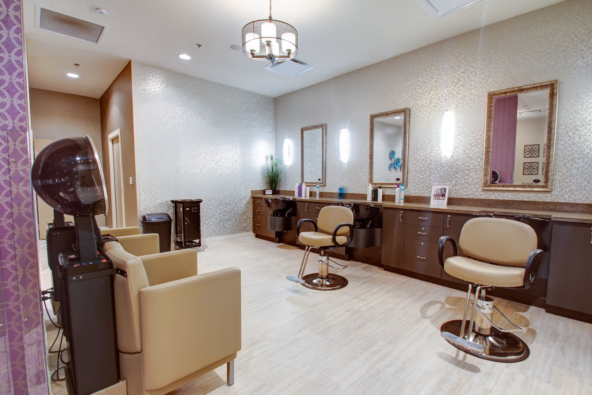 A modern salon area inside a senior living facility with beige salon chairs, hair washing stations, large mirrors on the wall, and a hair dryer hood. The room has light-colored flooring, patterned wallpaper, and soft lighting fixtures.