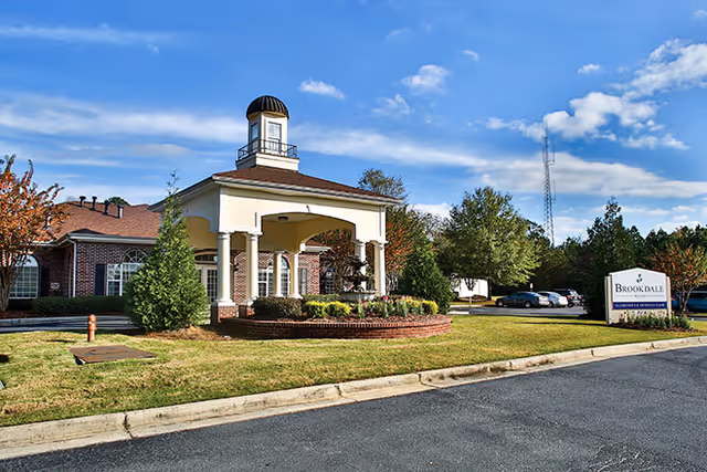 Exterior view of a senior living facility with a covered entrance featuring columns and a small tower structure on the roof. The building is surrounded by well-maintained landscaping including bushes and trees. A parking lot with several cars is visible in the background under a partly cloudy blue sky. A sign near the parking lot reads 'Brookdale'.