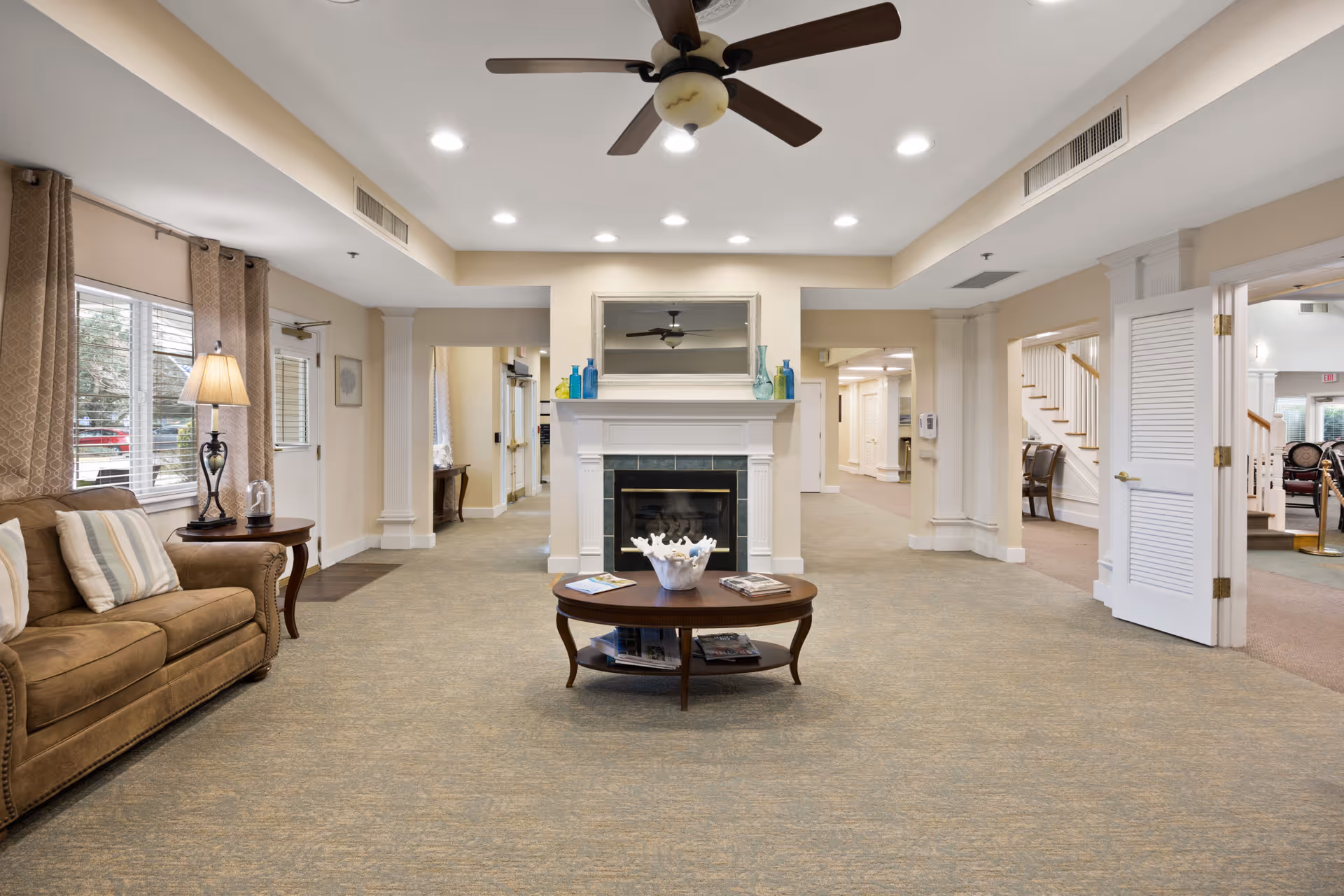 A spacious and well-lit common area with beige walls and carpeted floor. There is a brown sofa with striped pillows on the left side next to a window with curtains and a table lamp. In the center, there is a wooden coffee table with magazines and a decorative bowl, positioned in front of a white fireplace with a mirror and decorative vases on the mantel. Ceiling lights and a ceiling fan are visible above. Multiple doorways and hallways lead to other parts of the facility, including a staircase on the right side.