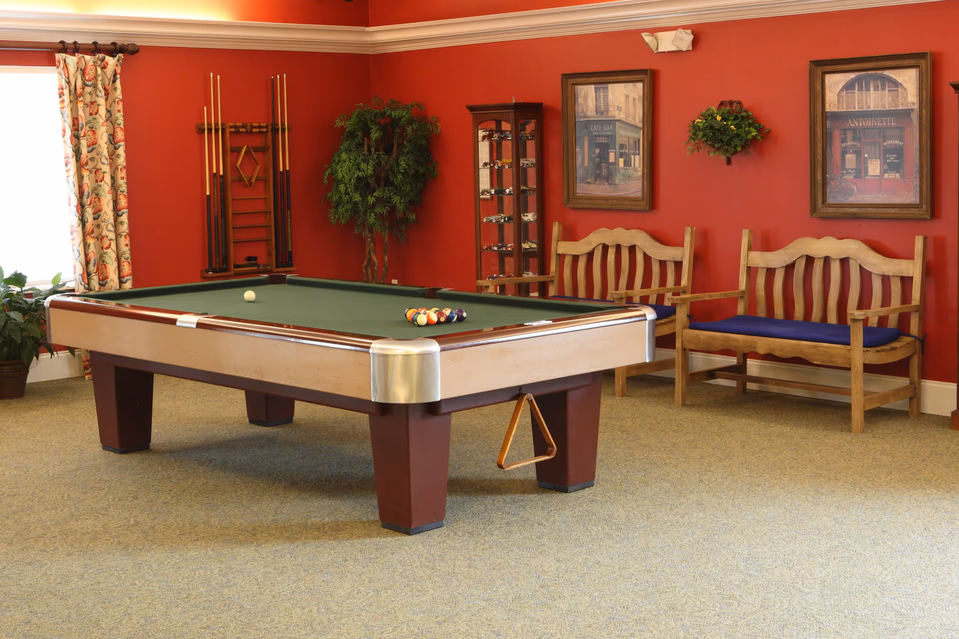 A recreational room with a pool table in the center, pool cues mounted on the wall, two wooden benches with blue cushions, framed artwork, a potted plant, and a red painted wall.