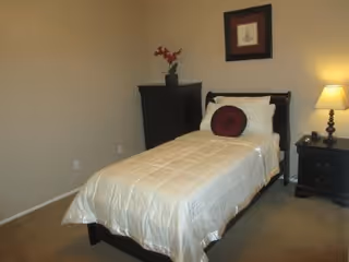 A small bedroom with a single bed covered in a cream-colored comforter and a round burgundy pillow. There is a dark wooden nightstand with a lamp and a few items on it to the right of the bed. A dark wooden dresser with a vase of red flowers is against the back wall. A framed picture hangs above the bed on a beige wall.