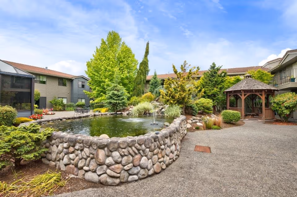 Outdoor courtyard area of a retirement and assisted living facility featuring a stone-bordered pond with a small fountain, surrounded by various green trees and shrubs. There is a wooden gazebo on the right side and multi-story residential buildings in the background under a partly cloudy blue sky.