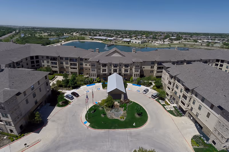 Aerial view of a U-shaped senior living complex surrounding a circular driveway and landscaped courtyard with a lake beyond.